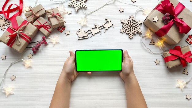 Criança segurando o smartphone com a posição horizontal de tela verde no fundo da mesa de madeira branca com presentes de natal. celular com chroma key. feche as mãos.