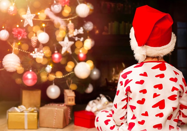 criança sentada em casa contra o fundo das luzes e presentes da árvore de Natal Vista traseira