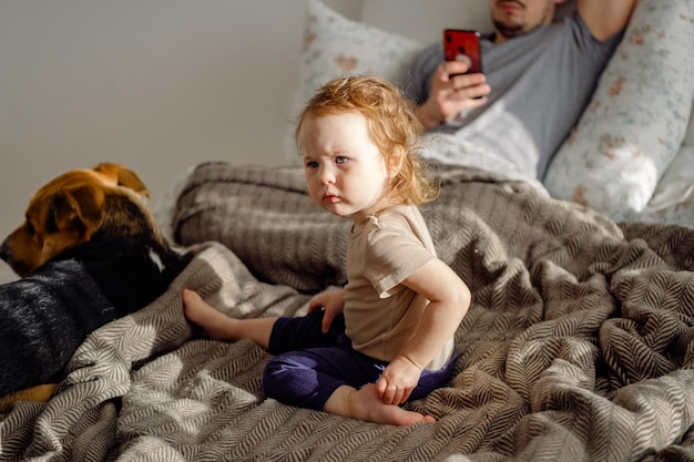 Criança sentada na cama com o pai usando celular e cachorro doméstico no fundo