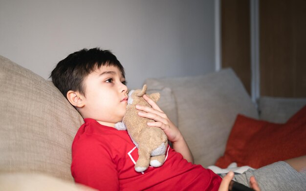 Criança sentada no sofá assistindo TV Um menino segurando o controle remoto e olhando com pensamentos profundos Criança positiva deitada no sofá gosta de assistir desenhos animados na TV Criança relaxando em casa no fim de semana