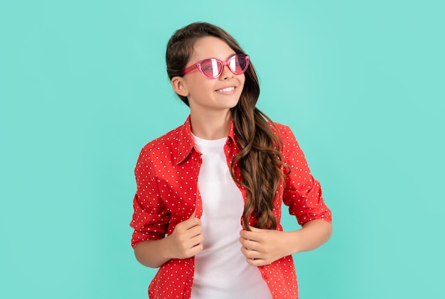 Criança sorridente, cabelo longo e encaracolado, posando em óculos de sol e estilo de moda de camisa casual