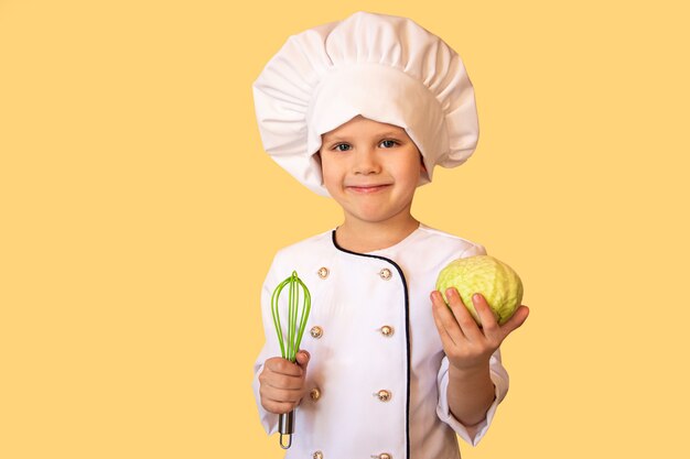 Criança sorridente com uniforme branco do chef, segurando um repolho e um batedor nas mãos. fundo amarelo alegre.