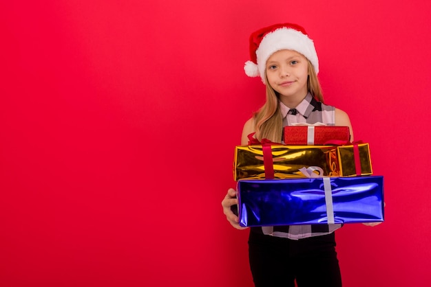 Criança sorridente e alegre com chapéu de Papai Noel segurando um presente de Natal na mão sobre a imagem de fundo vermelho