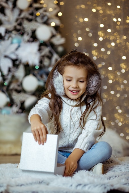 Criança sorridente feliz abrindo uma caixa de presente perto da árvore de natal decorada com luzes