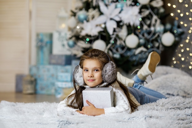 Criança sorridente segurando uma caixa de presente perto da árvore de natal decorada com luzes