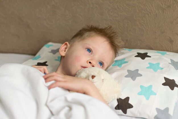 Criança triste doente com temperatura deitada na cama com um urso de brinquedo doença do vírus do resfriado da gripe