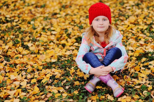 Foto criança - uma menina com um chapéu de malha vermelho sentado sobre um fundo amarelo folhas caídas de outono.