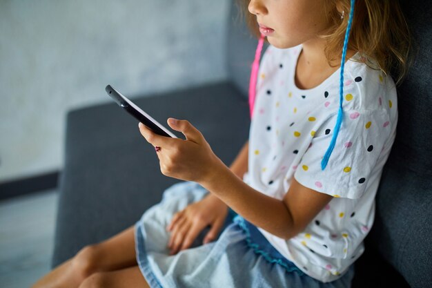 Criança usando celular no sofá em apartamento moderno e luminoso, menina brinca no smartphone em casa, problema de vício em gadgets