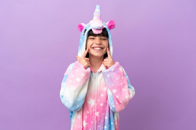 Criança vestindo um pijama de unicórnio isolado no fundo roxo sorrindo com uma expressão feliz e agradável