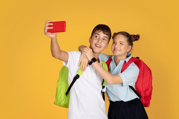 Crianças adolescentes europeias positivas com mochilas abraçando tirando selfie no smartphone na escola