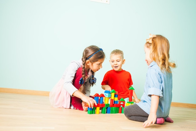 Crianças alegres brincando na creche. Três pré-escolares, duas meninas adoráveis e um menino sorridente e bonito, construindo uma cidade de brinquedo de cubos de madeira coloridos. Brinquedos educativos para pré-escola e jardim de infância.