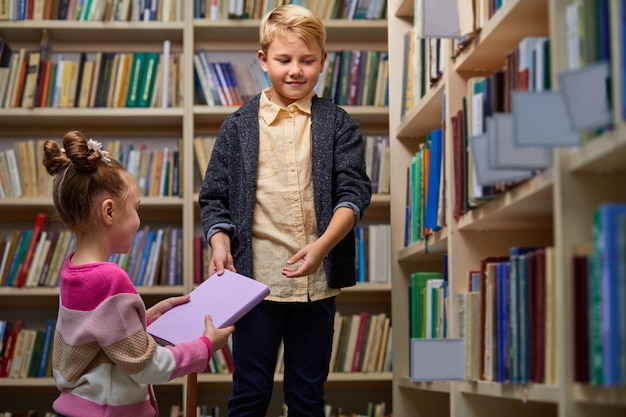 crianças amigáveis dando livros umas para as outras, ajudando na biblioteca antes da escola, sorrindo