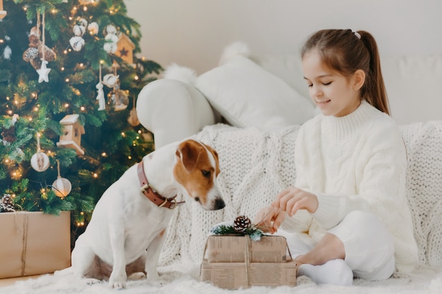 Crianças, animais e conceito de férias de inverno. Encantadora menina Europeia descompacta caixa de presente, coloca no chão, juntamente com o filhote