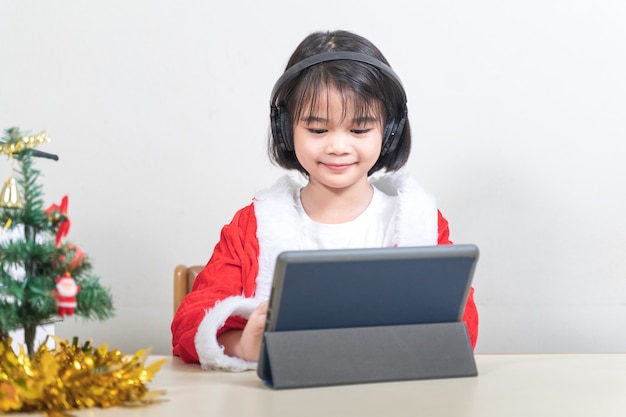 Crianças asiáticas menina vestem Natal Papai Noel conversando com amigos no tablet digital. Conceito de férias de Natal Foto de stock