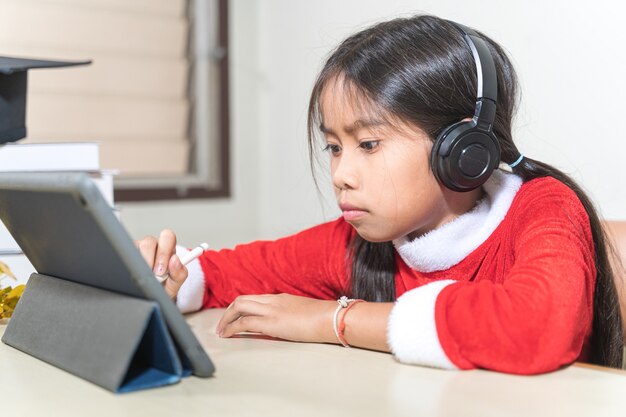 Crianças asiáticas menina vestem Natal Papai Noel conversando com amigos no tablet digital. Conceito de férias de Natal Foto de stock