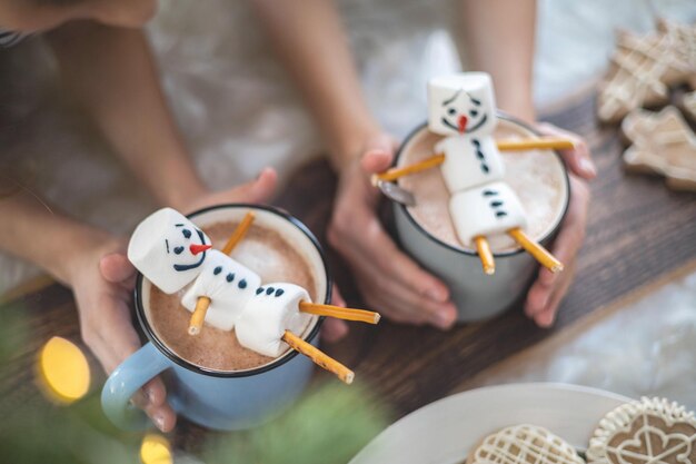 Crianças bonitas brincam com bonecos de neve de tricô e comem pão de gengibre e bebem cacau com marshmallows conceito de preparação de Natal
