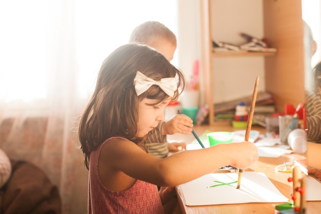 Crianças bonitas pintando com pincel e tintas coloridas Garota adorável de 4 anos desenhando em papel branco perto da janela na sala de luz