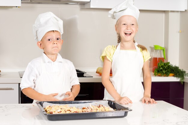 Crianças brancas em trajes de chefs fazem pizza saborosa na cozinha