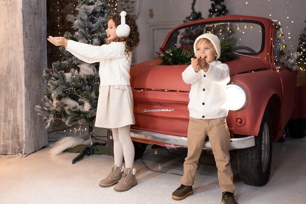 Crianças brincando com neve perto de um carro vermelho e uma árvore e luzes de natal