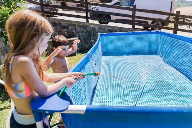 Crianças brincando em uma piscina em sua casa rural, enchendo a piscina com uma mangueira de água Crianças felizes brincando com água