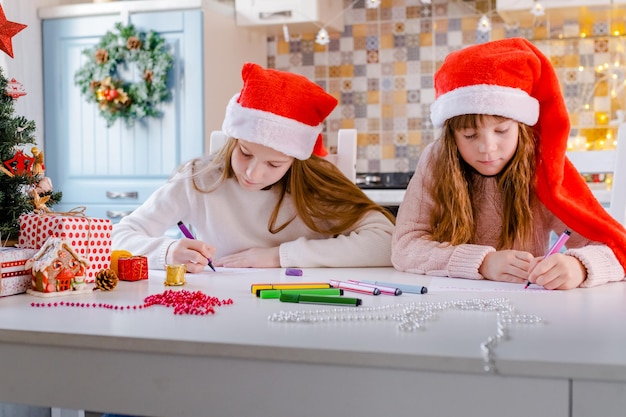 Crianças com gorros de papai noel escrevem uma carta para o papai noel na cozinha decorada de natal