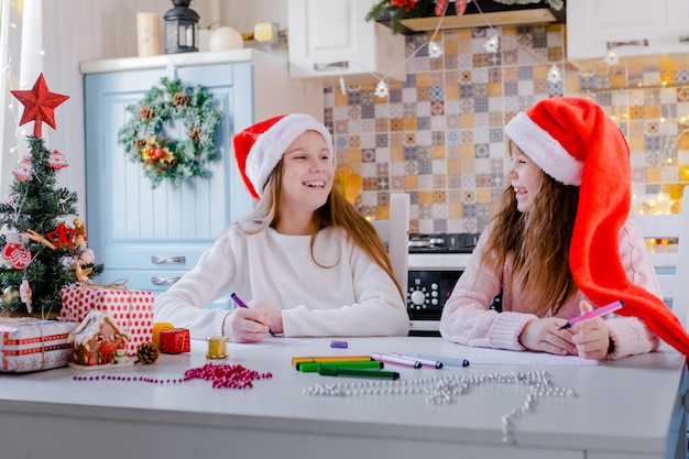 Crianças com gorros de papai noel escrevem uma carta para o papai noel na cozinha decorada de natal