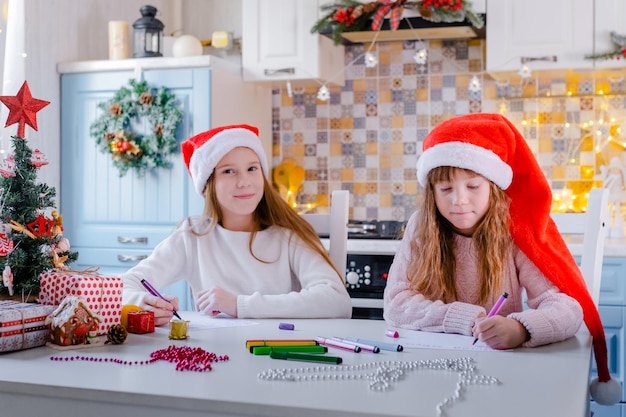 Crianças com gorros de papai noel escrevem uma carta para o papai noel na cozinha decorada de natal