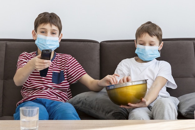 Foto crianças com máscaras médicas assistindo tv e comendo pipoca