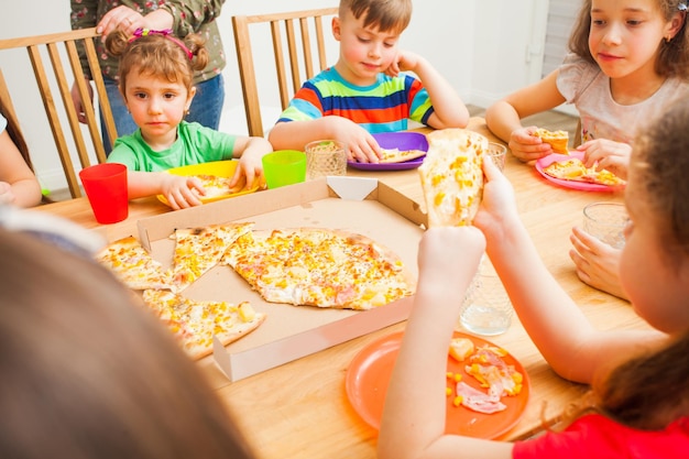 Crianças comendo pizza, mesa de madeira na sala, festa de amigos