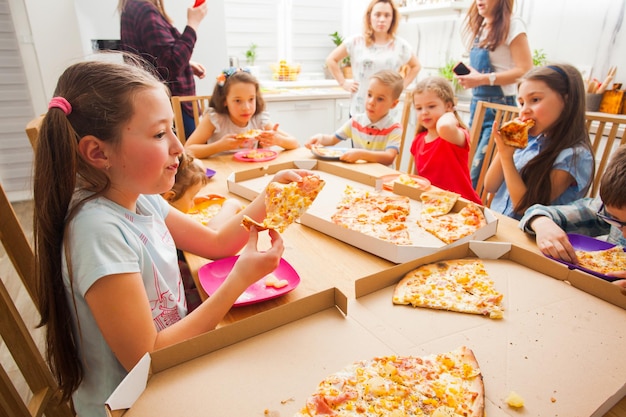 Crianças comendo pizza na mesa da cozinha e as mães estão conversando