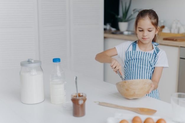 Crianças cozinhando e conceito de casa Menina de cabelos escuros bate ingredientes na tigela ocupada preparando massa para bolo sendo futuro chef usa camiseta branca e avental listrado usa ingredientes