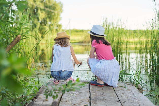 Crianças de duas garotas bonitas sentadas no cais de madeira do lago em juncos, brincando com água, conversando, vista traseira. Férias de verão, natureza, infância feliz, amizade, estilo country.