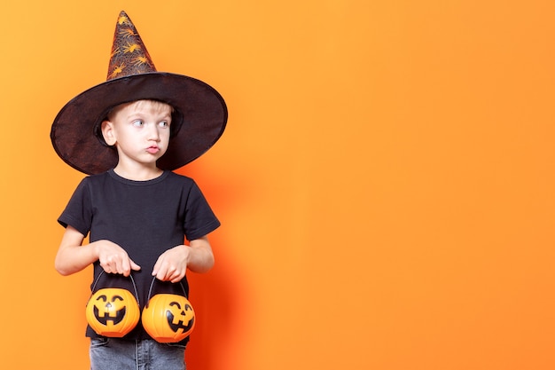 Crianças de Halloween. Menino com chapéu de feiticeiro, segurando uma cesta em forma de abóbora com doces em um fundo laranja, copie o espaço. A criança quer comer doce. Tradição doçura ou travessura de Halloween