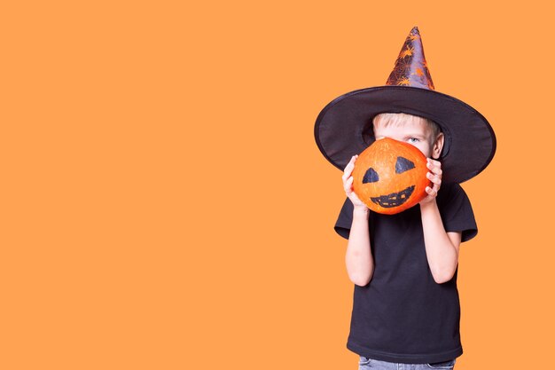 Crianças de Halloween. Um menino com um chapéu de mago segurando uma abóbora na frente do rosto com um rosto assustador pintado em um fundo laranja, copie o espaço. Humor da temporada de Halloween. Conceito feliz dia das bruxas