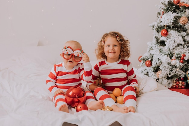 Crianças de pijama vermelho e branco experimentam óculos engraçados com papai noel sentado na cama. estilo de vida. irmão e irmã comemorando o natal. menino e menina estão jogando em casa. foto de alta qualidade