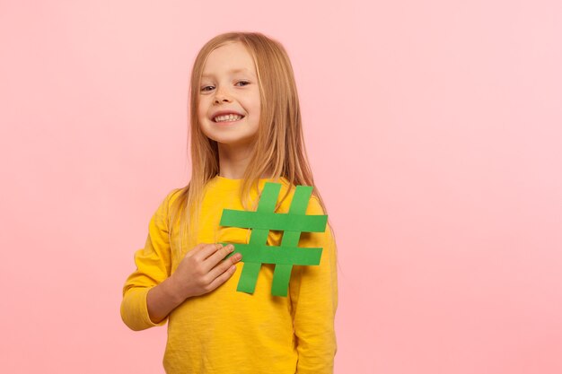 Crianças e popularidade na internet. retrato de uma menina linda feliz segurando o símbolo da hashtag e olhando para a câmera com um sorriso, mostrando o sinal de hashtag. foto de estúdio isolada em fundo rosa