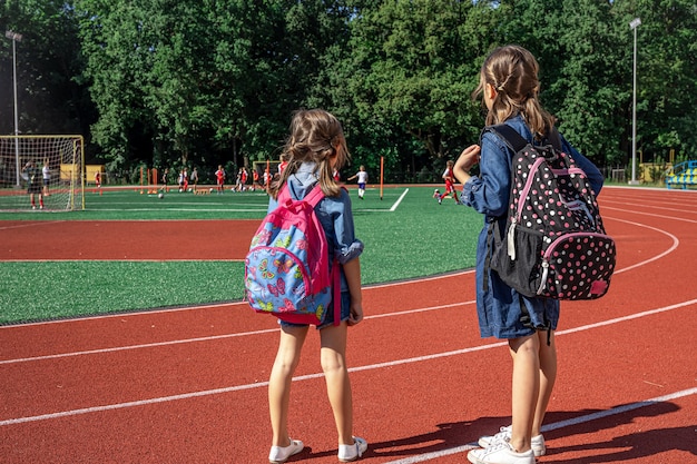 Crianças em idade escolar com mochilas no estádio, vendo os meninos jogarem futebol.