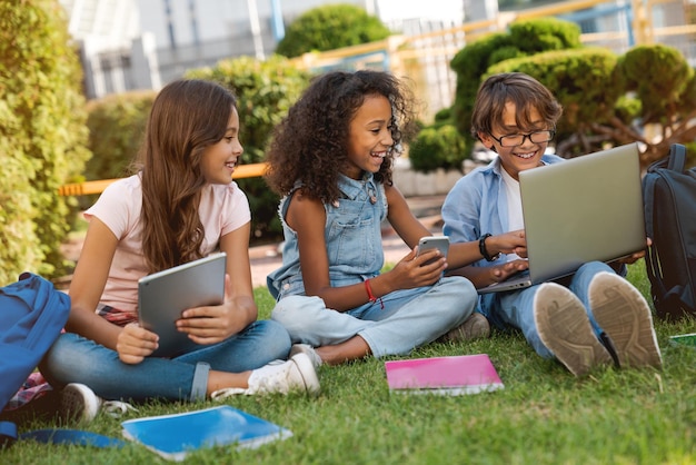 Crianças em idade escolar felizes com aparelhos digitais e mochilas sentadas no parque de grama