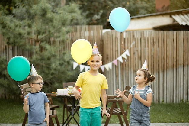 Crianças engraçadas e fofas comemorando aniversário com a família ou amigos em uma festa de aniversário no quintal Garoto usando chapéu de festa e lançando balões no céu