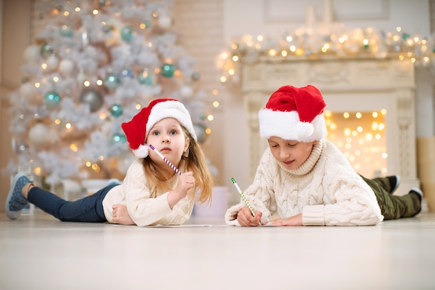 Foto crianças escrevendo uma lista de desejos no chão com uma decoração de natal no fundo