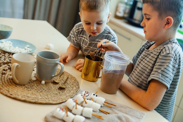 Crianças fazem bonecos de neve com marshmallows e canudos salgados bebem cacau na cozinha de casa preparando ...
