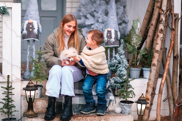 Crianças felizes com cachorrinho branco sentado na varanda da casa decorada de Natal, nevando ao ar livre. Feliz Ano Novo e feliz Natal. Inverno mágico