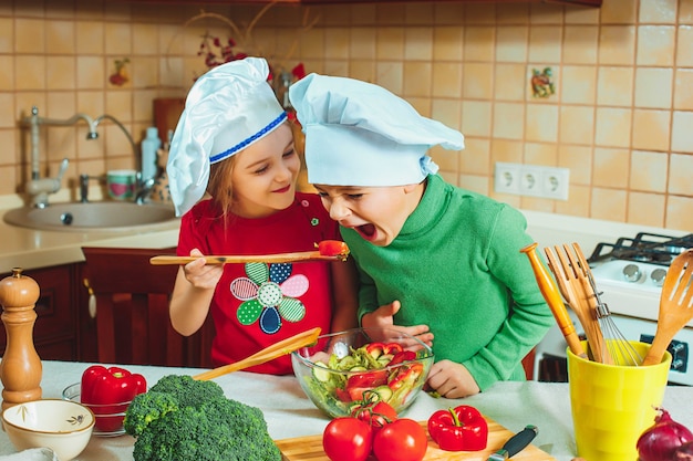 Crianças felizes e engraçadas da família estão preparando uma salada de legumes fresca na cozinha