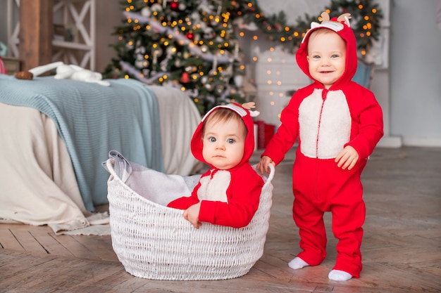 Crianças gêmeas em ternos vermelhos de Papai Noel estão sentados lado a lado em casa contra o fundo de uma árvore de Natal