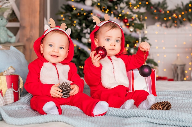 Crianças gêmeas em ternos vermelhos de Papai Noel estão sentados lado a lado em casa contra o fundo de uma árvore de Natal