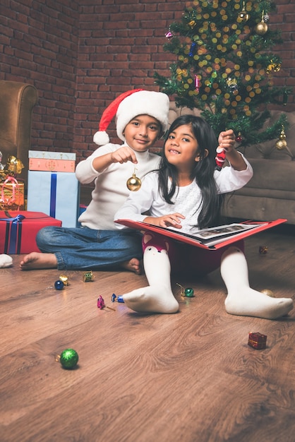 Foto crianças indianas asiáticas fofas celebrando o natal em casa com o chapéu do papai noel, presentes e árvore de natal