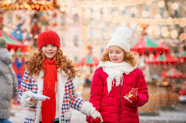 Crianças, irmãs ruivas aquecem as mãos enluvadas com uma caneca de chá quente em um mercado de Natal festivamente decorado na cidade.