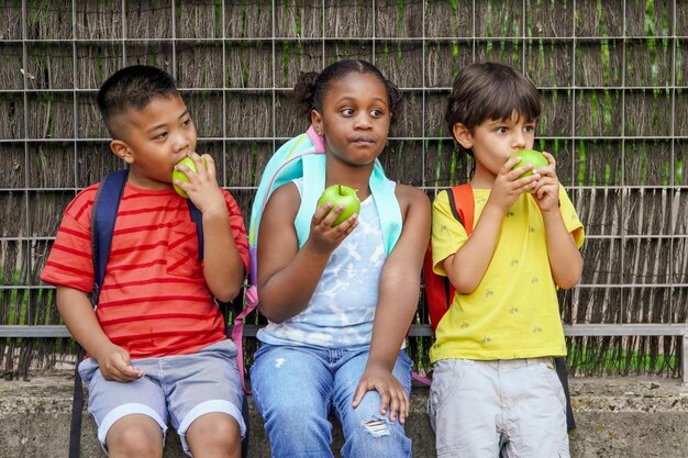 Crianças multiétnicas com mochilas comendo maçãs na entrada da escola