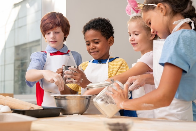 Crianças multiétnicas de aventais olhando umas para as outras enquanto discutem o cardápio festivo enquanto fazem biscoitos