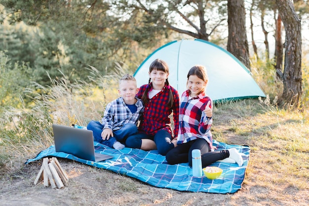 Crianças olhando show no laptop na floresta Meninos e meninas estão descansando em um acampamento As crianças sentam-se na manta na grama no verão